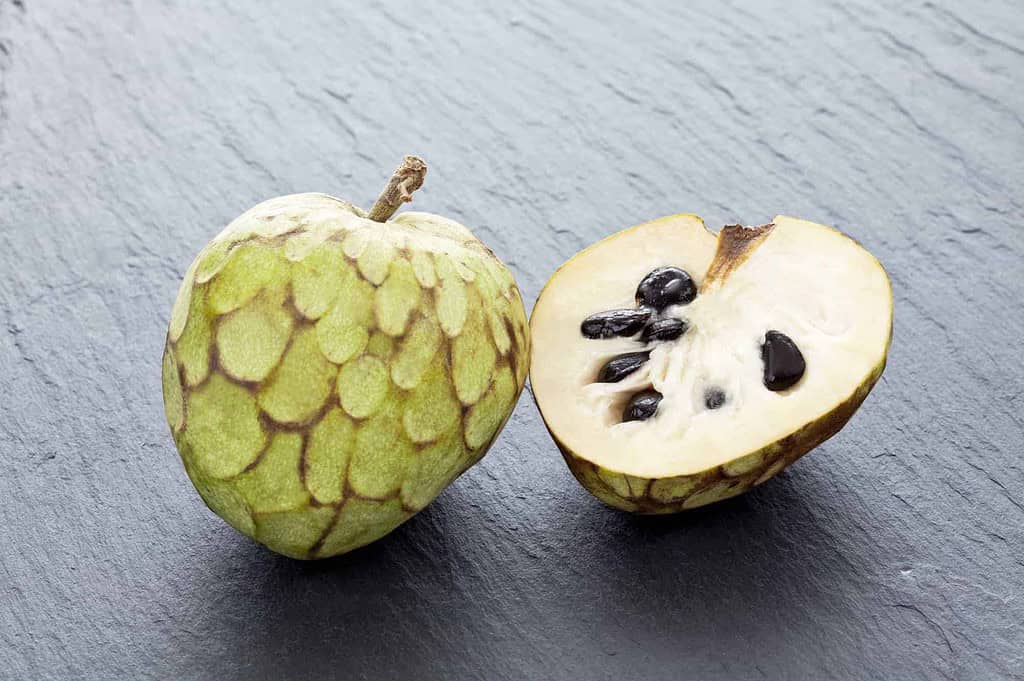 A cherimoya fruit displayed on a dark slate surface, with one half cut open to reveal its creamy white flesh and glossy black seeds. The uncut half showcases its scaly green exterior