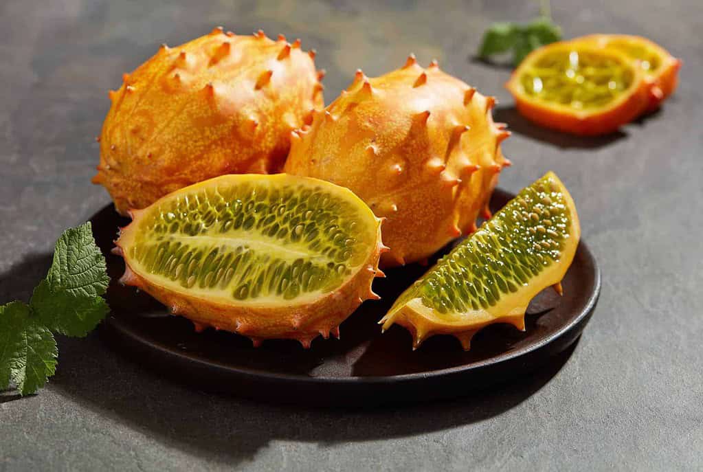 Bright orange kiwano (horned melon) fruits on a dark plate, with one cut open to reveal its vibrant green, jelly-like flesh filled with seeds. A few fresh mint leaves and additional fruit slices are placed in the background for an elegant presentation.