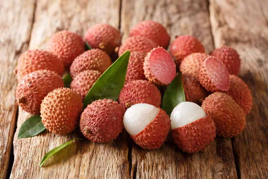 A pile of fresh lychee fruits on a rustic wooden surface, with some peeled to reveal their smooth, translucent white flesh. Green leaves are scattered among the vibrant red fruits, adding a natural touch.