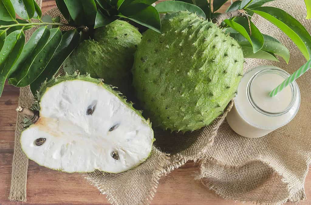 Fresh soursop fruits placed on a rustic wooden table with one cut open, revealing its white, fibrous flesh and black seeds. The setup includes green leaves and a glass of soursop juice with a straw, presented on a burlap cloth for a natural, tropical vibe