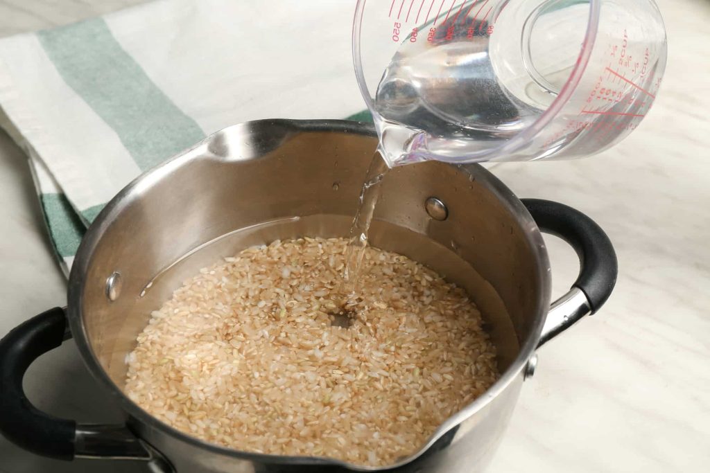 Water is being poured from a measuring cup into a stainless steel pot containing uncooked brown rice. The pot, equipped with black handles, is placed on a light-colored countertop next to a neatly folded green-and-white kitchen towel. The clear measuring cup displays red measurement markings, emphasizing the importance of precision in the cooking process. 
