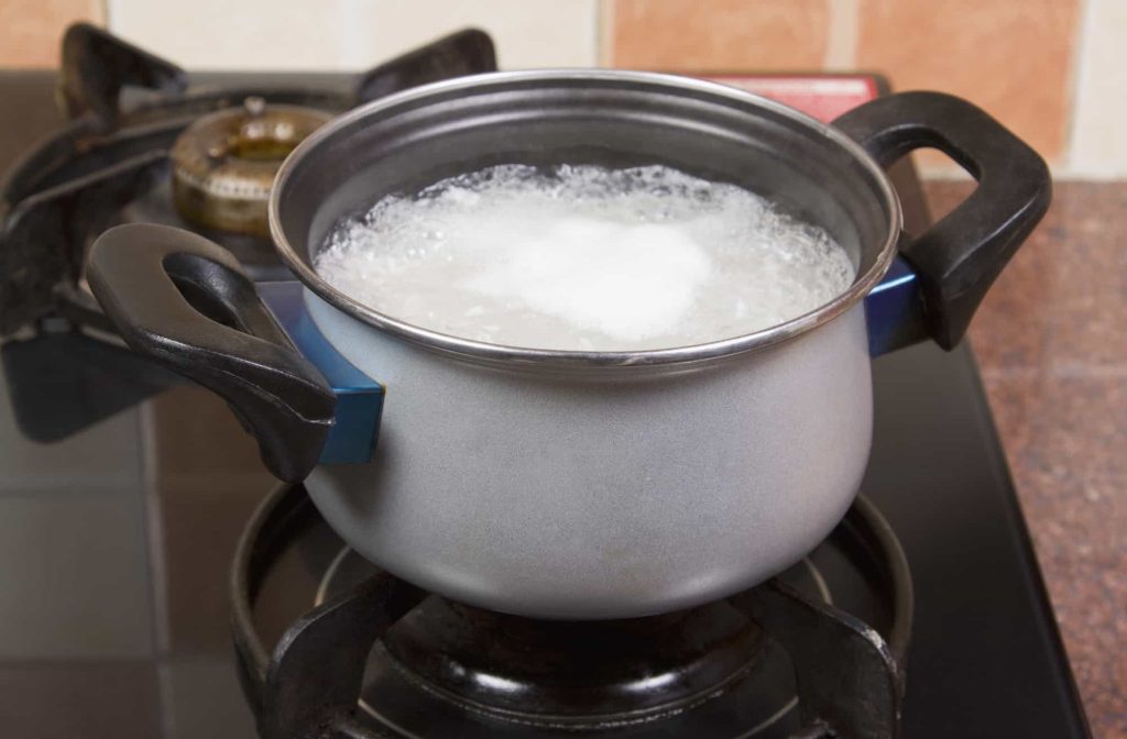 A small gray pot with black handles sits on a gas stove, filled with boiling water and rice. The bubbling foam at the surface indicates the rice is actively cooking. The kitchen background includes tiled walls and additional stove components, creating a cozy and practical cooking environment. 