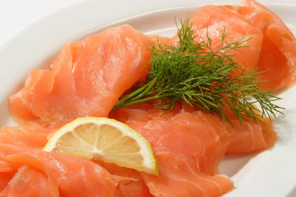 Slices of smoked salmon, featuring a crispy touch of salmon skin, are garnished with a sprig of dill and a lemon wedge on a white plate.