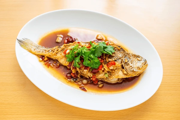 A whole fried croaker fish served on a white oval plate, topped with fresh cilantro, sliced red chilies, and a savory sauce with garlic and soy-based flavors. The dish is presented on a light wooden table for a simple and appetizing display