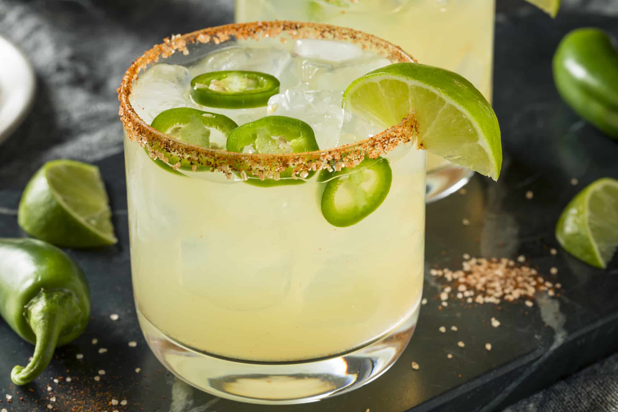 A glass of cocktail with ice and sliced jalapeños, garnished with a chili powder rim and a lime wedge, showcases the vibrant zest of Jalapeno Juice. More lime slices and jalapeños are in the background on a dark surface, tempting you to learn how to make this spicy drink.