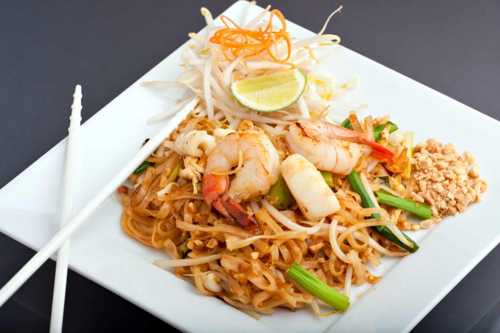 Seafood pad Thai dish of Thai fried vermicelli rice noodles on a square white plate with chopsticks and grated carrot garnish.