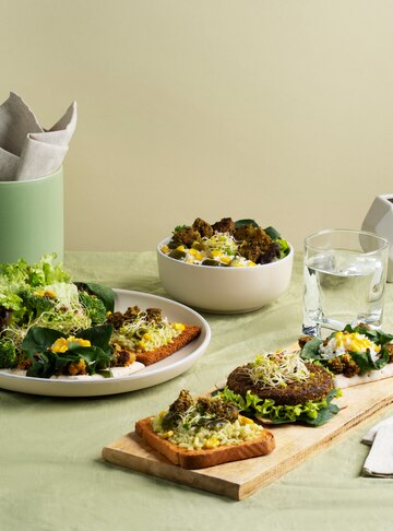 A plant-based meal arrangement featuring a wooden board with avocado toast, leafy greens, sprouts, and a plant-based burger patty. Surrounding it are a plate with a fresh green salad and a bowl of mixed salad with corn and leafy greens. A glass of water and a napkin holder complete the minimalist table setting on a light green tablecloth. 