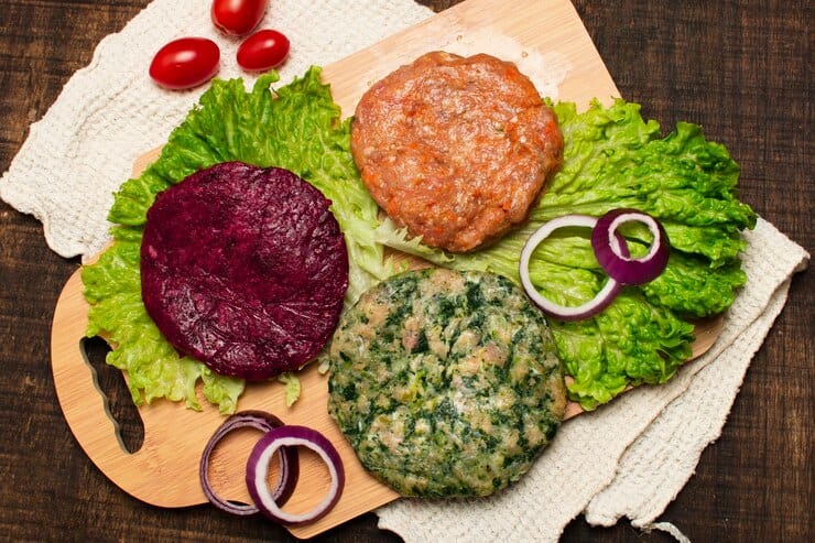 The image shows a wooden board with three colorful plant-based burger patties placed on fresh lettuce leaves. The patties vary in color and composition: one is red (likely beet-based), one is green (possibly made with spinach or herbs), and one is a natural brown hue (resembling a classic plant-based meat patty). The board is garnished with red onion slices and small cherry tomatoes, creating a vibrant and appetizing display. 