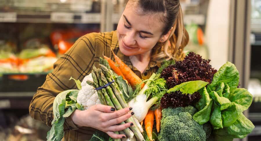 In front of a refrigerator, a woman holds a vibrant assortment of vegetables, highlighting her commitment to vegan grocery shopping.
