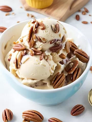 A blue bowl brims with two scoops of vanilla ice cream crowned with pecans, evoking the delight of an Almond Joy Sundae. Additional pecans are scattered around the table, and a cutting board provides a rustic background.