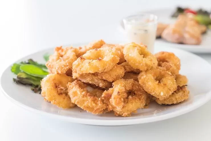 A plate of golden brown, crispy fried popcorn shrimp sits enticingly, accompanied by a small cup of dipping sauce. This delectable seafood delight is garnished with fresh green lettuce on the side, perfectly capturing ocean flavors in every bite.