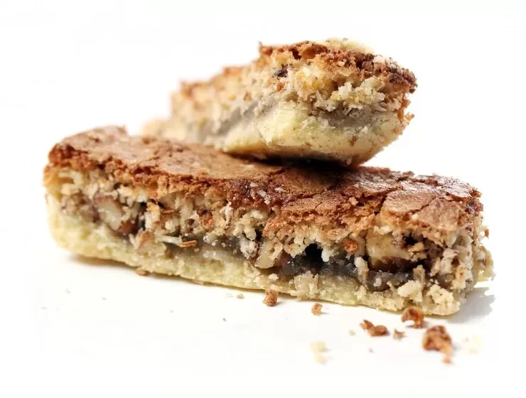 Two delicious paleo pecan pie bars stacked on a white surface. The bars boast a crumbly crust with a visible layer of chopped pecans and a golden-brown top. Crumbs are scattered around them, offering a tempting nod to indulgence while staying true to classic flavors.