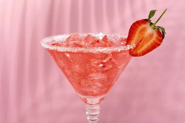 A paleo strawberry daiquiri in a martini glass, filled with ice and garnished with a halved strawberry on the rim. The sugared rim complements the fresh berry, while the background is a matching pink shade.