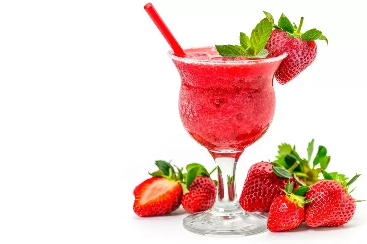 A paleo strawberry daiquiri in a glass with a sugared rim, garnished with a whole strawberry and mint leaves. A red straw is in the drink. Fresh strawberries are scattered around the base of the glass against a white background.