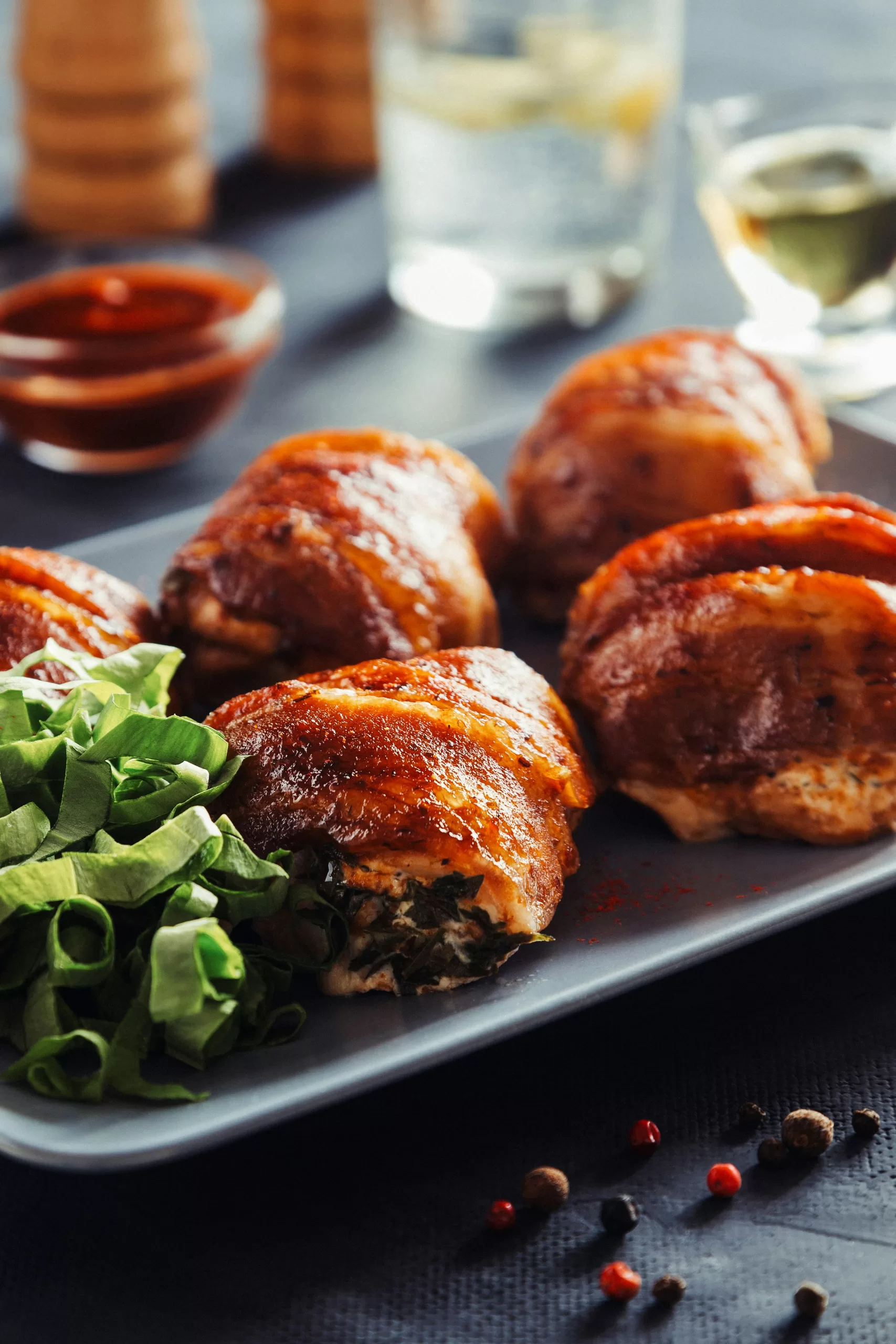 A plate of paleo-friendly, pizza-stuffed chicken rolls is garnished with a side of fresh greens. In the background, pepper mills, a glass of water, and a small bowl of sauce complete the scene. Peppercorns are scattered on the table.