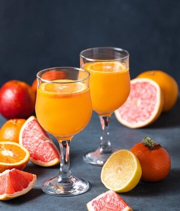 Two glasses of orange juice, topped with vibrant orange slices, sit amidst a cheat sheet of citrus fruits—whole and sliced oranges, grapefruits, and a lemon—all set against a dark background. Perfect for anyone drinking in the beauty of Paleo drinks.