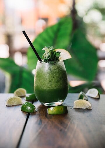 A refreshing green smoothie with a black straw, garnished with a lemon slice and mint, sits on a wooden table surrounded by lime wedges. Embracing this paleo drink, the scene is complemented by a blurred large green leaf in the background.