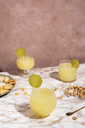 Three glasses of a refreshing yellow paleo drink garnished with lime slices sit on a marbled table, perfect for a paleo cheat sheet gathering. A plate of crackers and cheese rests on the left, while scattered pistachios and a spoon occupy the right. The background is a textured brown wall.