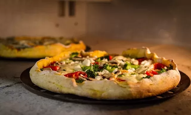 Close-up of two homemade paleo pizzas, including a gluten-free option, baking in an oven. The foreground pizza features toppings like cheese, mushrooms, red bell peppers, and greens on a golden crust. The background pizza is slightly out of focus. The scene is warmly lit.