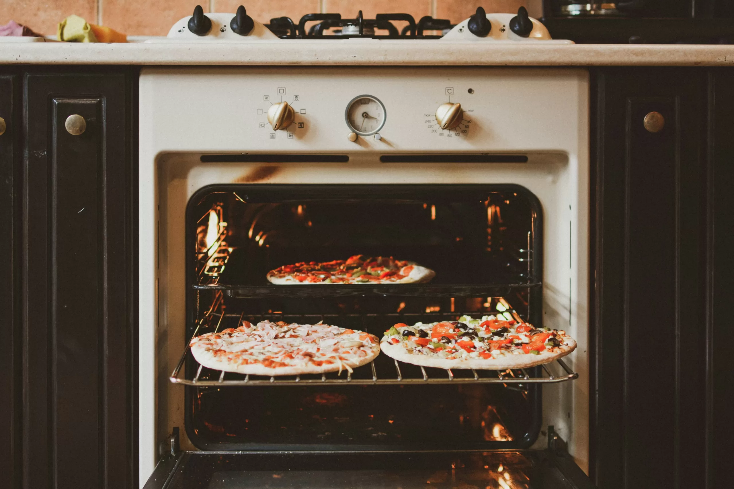 An oven with its door open reveals two racks of gluten-free paleo pizzas baking inside, each topped with various ingredients. The kitchen boasts a traditional design with dark cabinets and a stove above the oven, offering a cozy culinary haven.