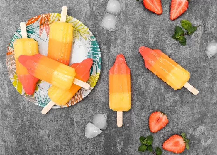 Colorful fruit paleo candy corn popsicles, perfect for a paleo treat, are arranged on a decorative plate against a gray background. More popsicles are scattered around with ice cubes, fresh strawberry halves, and leaves, creating a delightful visual feast.