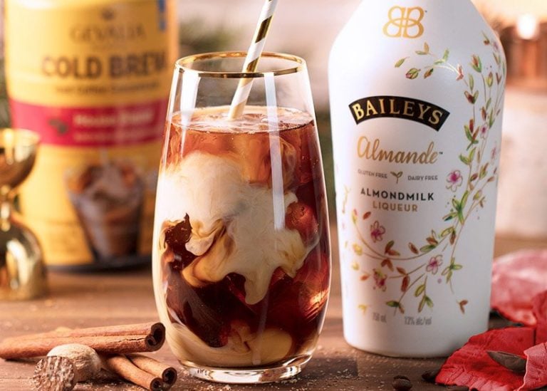 A glass of iced coffee swirls with almond milk liqueur next to a bottle of Baileys Almande. In the background, there’s a container of cold brew coffee, cinnamon sticks, and autumn leaves, creating a cozy fall atmosphere.