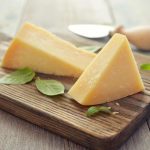 Two wedges of hard cheese on a wooden cutting board, garnished with fresh basil leaves. A cheese knife with a wooden handle is in the background on a rustic wooden surface, offering a healthy pairing for your culinary creations.