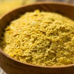 A wooden bowl filled with yellow nutritional yeast flakes sits on a wooden surface. The light, crumbly flakes highlight the yeast's textured surface, showcasing its essential role in vegan cooking.