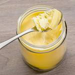 A jar of ghee with a metal spoon resting on top, containing a scoop of ghee. Perfect for baking or as a vegan butter alternative, the jar is placed on a wooden surface, and the ghee appears smooth and creamy.