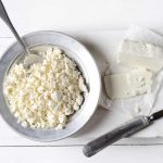 A bowl of crumbled Paleo-friendly white cheese with a spoon sits on a metal plate to the left. To the right, partially sliced cottage cheese rests on a cutting board with a knife beside it, all elegantly displayed on a white wooden surface.