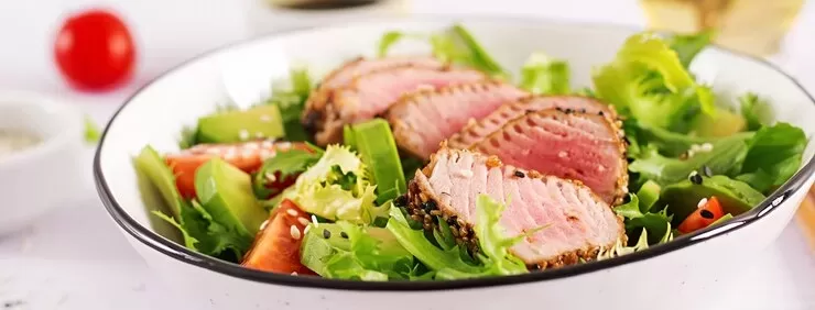 A fresh salad featuring sliced seared tuna fish, mixed greens, avocado, and cherry tomatoes, garnished with sesame seeds, served in a white bowl.