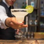 A bartender expertly serves a margarita in a salt-rimmed glass, perfectly garnished with a lime slice. The background is blurred, revealing shelves stocked with various bottles of alcohol, capturing the vibrant allure of cocktail artistry.