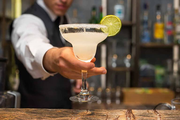 A bartender expertly serves a margarita in a salt-rimmed glass, perfectly garnished with a lime slice. The background is blurred, revealing shelves stocked with various bottles of alcohol, capturing the vibrant allure of cocktail artistry.