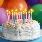 A white frosted cake with colorful sprinkles is topped with ten lit candles in blue and pink. Be careful not to eat candle wax as it glistens invitingly. The cake sits on a white plate, surrounded by colorful balloons in a festive setting.
