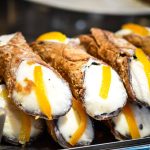 A close-up of several cannoli filled with creamy white ricotta, topped with slices of candied orange peel. The shells and filling create a delightful contrast as they are stacked neatly on a tray, showcasing their crispy, textured allure.