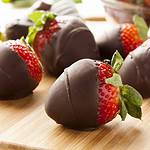 Close-up of several ripe strawberries dipped in dark chocolate, arranged on a wooden surface. In the background, a red rose is partially visible, adding to the image's romantic ambiance. For those wondering how long until the chocolate hardens, this serene timing guide invites patience.