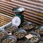 Metal bowls filled with clams sit on a market stall table, each brimming like a bushel of treasures. A scale is nestled among them, and seafood trays linger in the background. A handwritten price tag reads "20," reflecting the costs against a corrugated metal backdrop.
