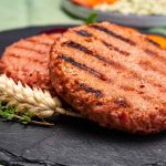 Two grilled plant-based burger patties rest on a slate plate, garnished with sprigs of thyme and wheat. The patties boast distinct grill marks and a textured surface, showcasing the appeal of vegan meat. A blurred background hints at a fresh salad to complement this delightful dish.