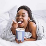 A person lying on a bed with braces smiles while eating cereal from a blue bowl. The striped blanket adds charm to the bright and airy room, creating a cozy and relaxed atmosphere.