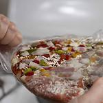 A person is unwrapping a frozen pizza topped with cheese, red and green peppers, and red onions. Though it arrives raw, it's ready for the oven beneath its clear plastic cover.