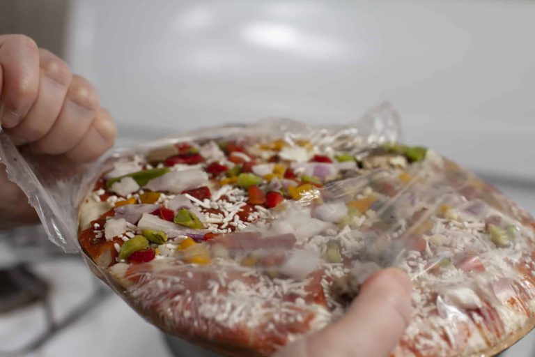 A person is unwrapping a frozen pizza topped with cheese, red and green peppers, and red onions. Though it arrives raw, it's ready for the oven beneath its clear plastic cover.