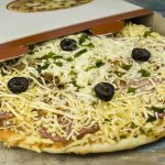 A partially open pizza box reveals a raw, uncooked pizza topped with shredded cheese, black olives, and herbs on a wooden surface.