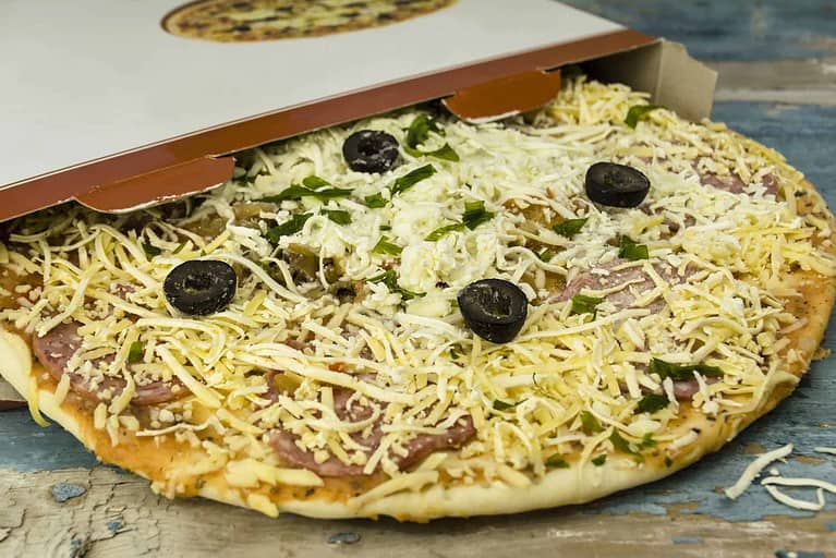 A partially open pizza box reveals a raw, uncooked pizza topped with shredded cheese, black olives, and herbs on a wooden surface.