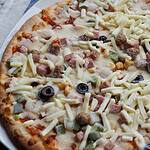 A close-up of a precooked frozen pizza topped with shredded cheese, olives, green and red peppers, sausage, and ham pieces on a thin crust, ready for baking.