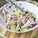 A decorative bowl on a wooden board contains sliced radishes, shredded cabbage, and a spoonful of dressing, with a spoon resting inside. The vibrant vegetables and dressing create a sushi-inspired salad appearance.