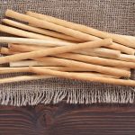 A pile of thin, crispy old breadsticks rests on a piece of burlap fabric. The burlap sits on a dark wooden surface, with the breadsticks arranged in a scattered manner.