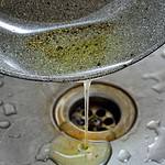 A pan with greasy olive oil residue is being poured into a metal sink drain, risking a clog. Water droplets are scattered around the sink.