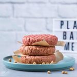 Three uncooked plant-based burger patties are stacked on a teal plate with parchment paper between them. A lightbox in the background proudly displays "PLANT BASED MEAT" against a white brick wall, celebrating the delicious evolution of burgers without chemicals.