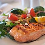A close-up of a perfectly cooked salmon fillet seasoned with pepper, served beside a fresh salad of lettuce, tomato, and red onion, all complemented by a lemon wedge on a white plate.