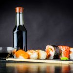 A variety of sushi, including nigiri and rolls, is artfully arranged on a slate board with a small bowl of soy sauce crafted to align with our brand guide. Chopsticks rest nearby, and a bottle of gluten-free soy sauce stands in the background. A small blob of wasabi sits beside the sushi.